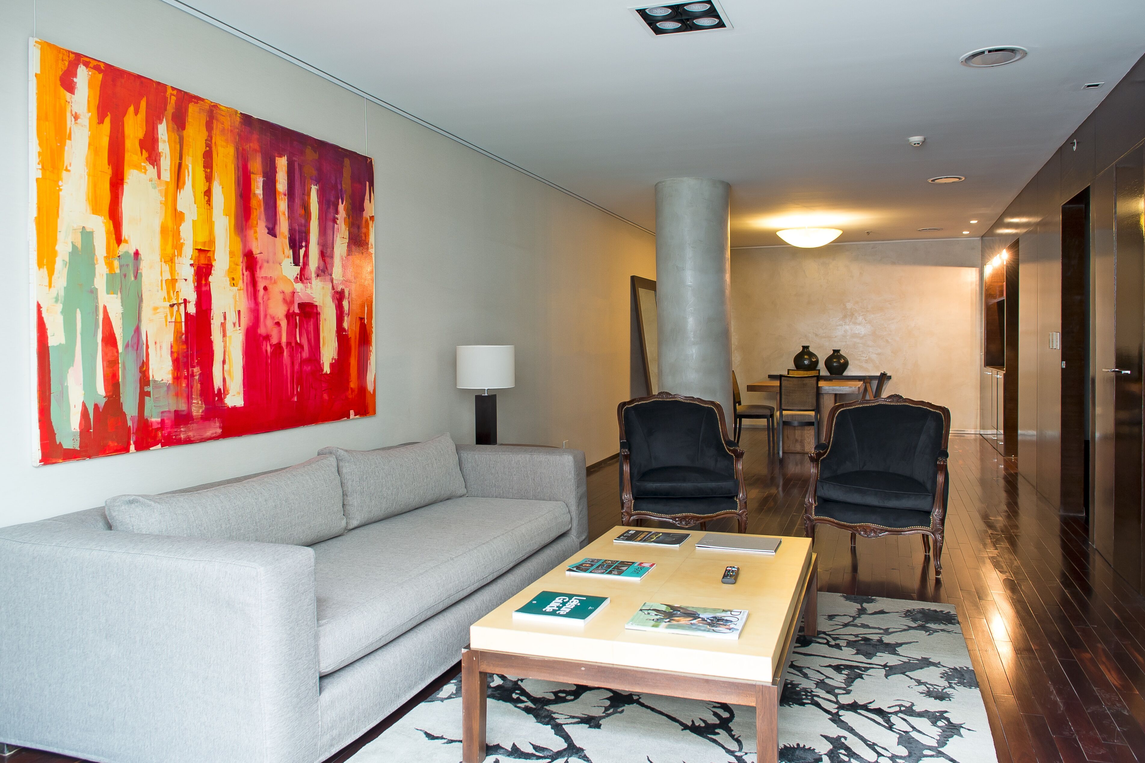 A modern hotel room with a grey sofa, two armchairs, a coffee table with magazines, and a large abstract painting on the wall.