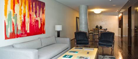 A modern hotel room with a grey sofa, two armchairs, a coffee table with magazines, and a large abstract painting on the wall.
