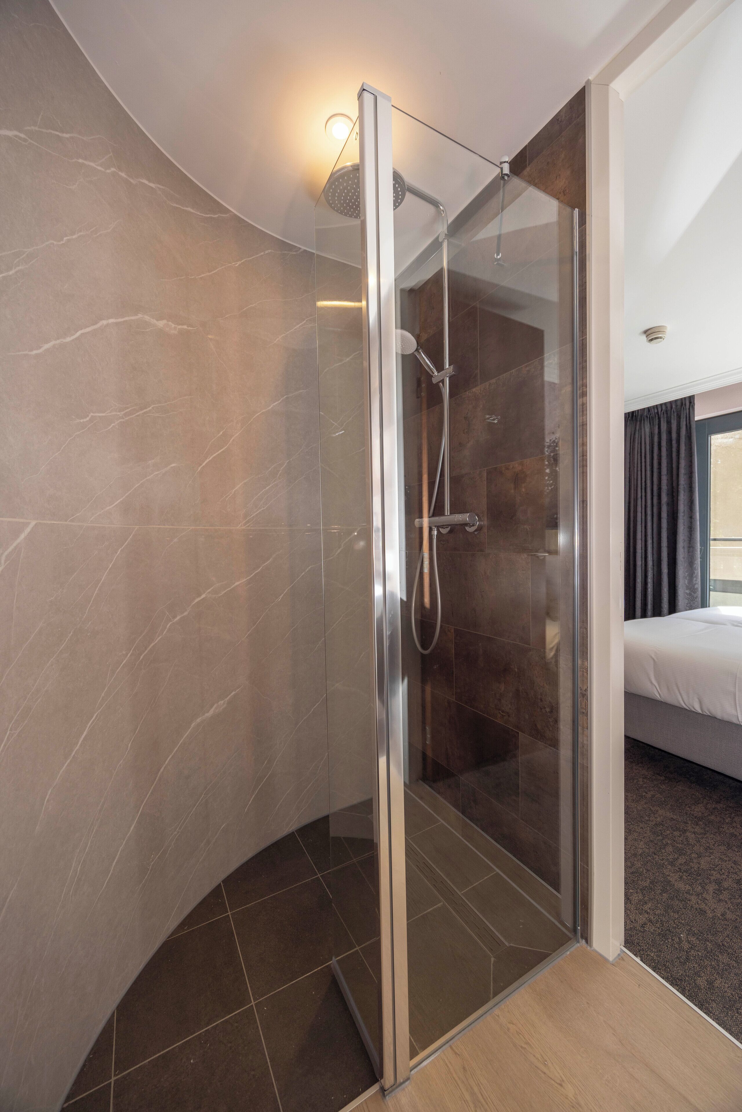 A modern bathroom with a glass-enclosed shower, a curved marble wall, and a visible bed in the adjacent room.