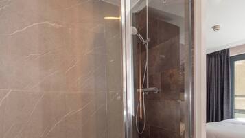 A modern bathroom with a glass-enclosed shower, a curved marble wall, and a visible bed in the adjacent room.