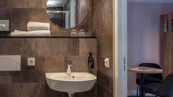 A modern bathroom with a white sink, mirror, and toilet. There is a small trash bin and a towel rack with towels.