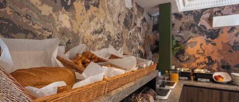 A breakfast buffet with wicker baskets of bread and pastries, a fruit bowl, and a juice dispenser.