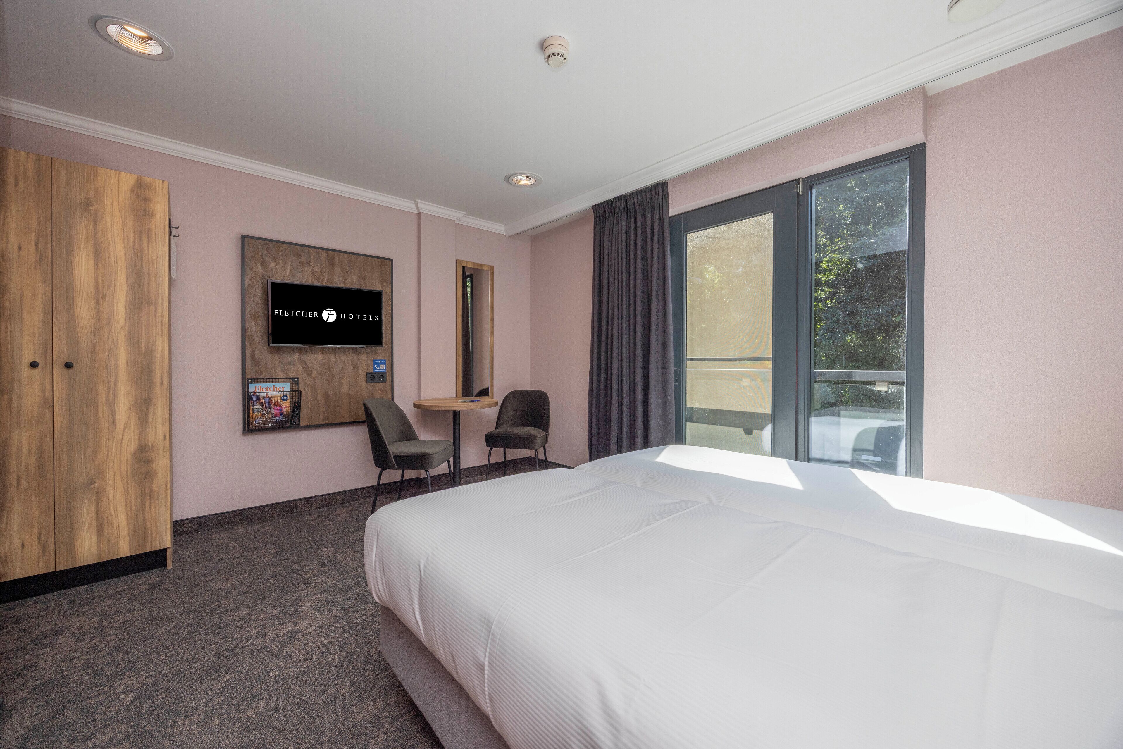 A hotel room with a bed, a desk, two chairs, a television, and a window.