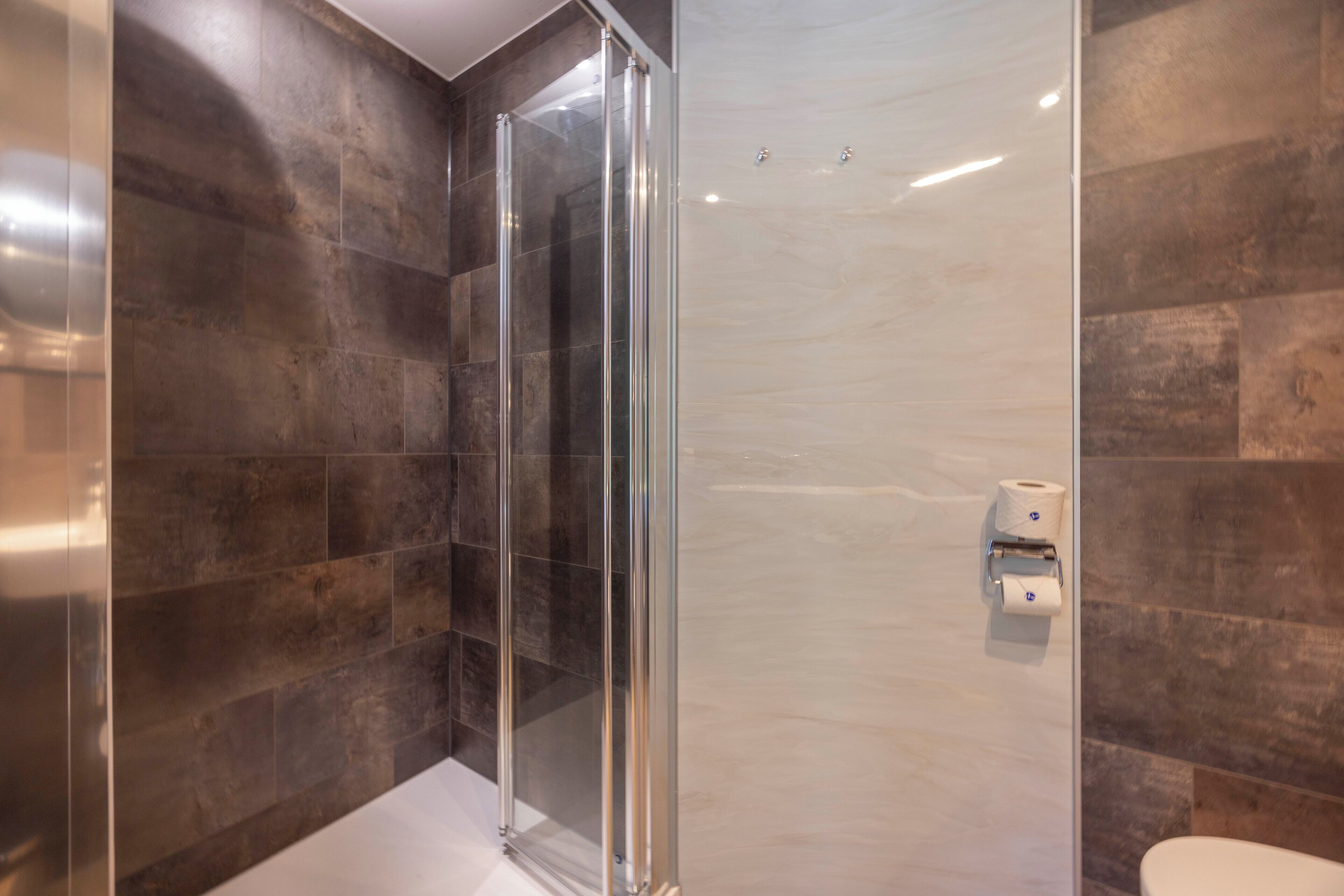 Une salle de bain moderne avec une douche à parois de verre, des murs recouverts de carreaux foncés et un porte-rouleau de papier toilette.