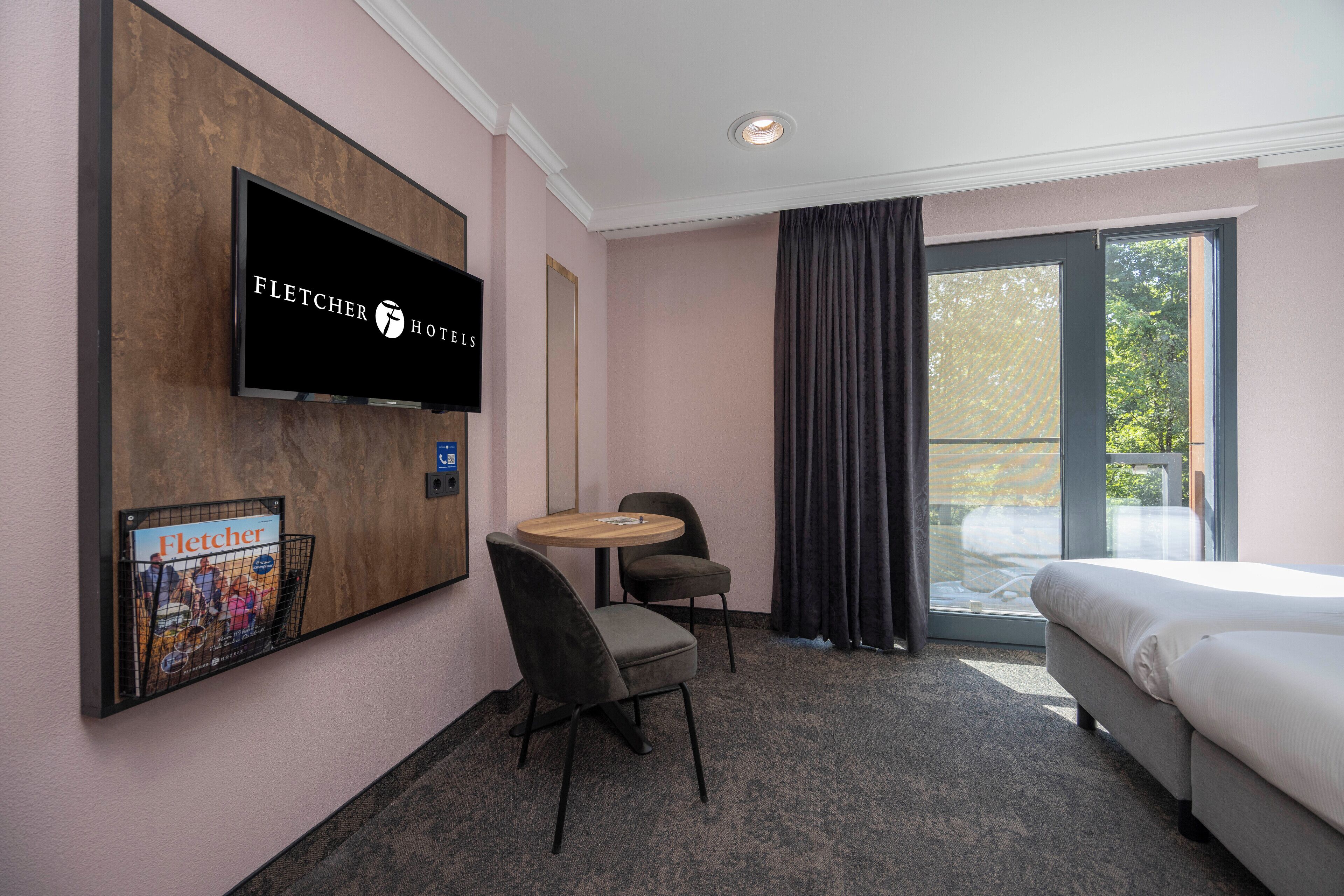 Habitación de hotel con cama, televisor montado en la pared, una mesita redonda, dos sillas y un ventanal con cortinas.
