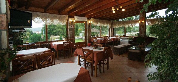 A restaurant with wooden tables and chairs, a TV mounted on the wall, and large windows offering a view of the outdoors.