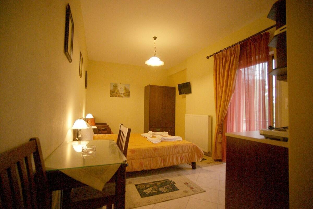Quarto pequeno com uma cama de solteiro, uma mesinha e uma cadeira. Há um radiador na parede e uma janela com cortinas.