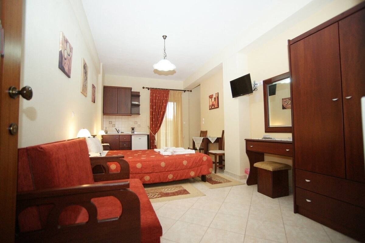 Quarto de hotel compacto com cama, escrivaninha, guarda-roupa, televisão e cozinha compacta.