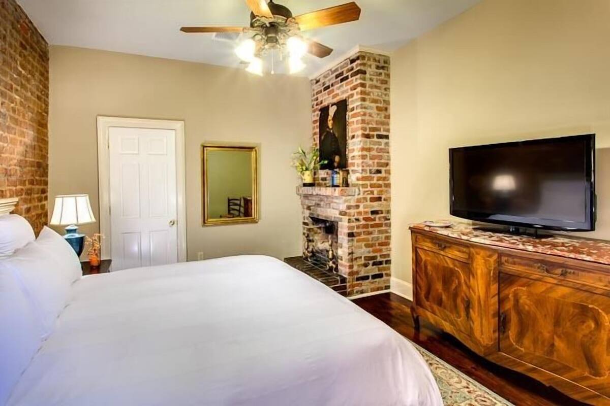 A bedroom with a brick fireplace, a bed, a television, and a ceiling fan.