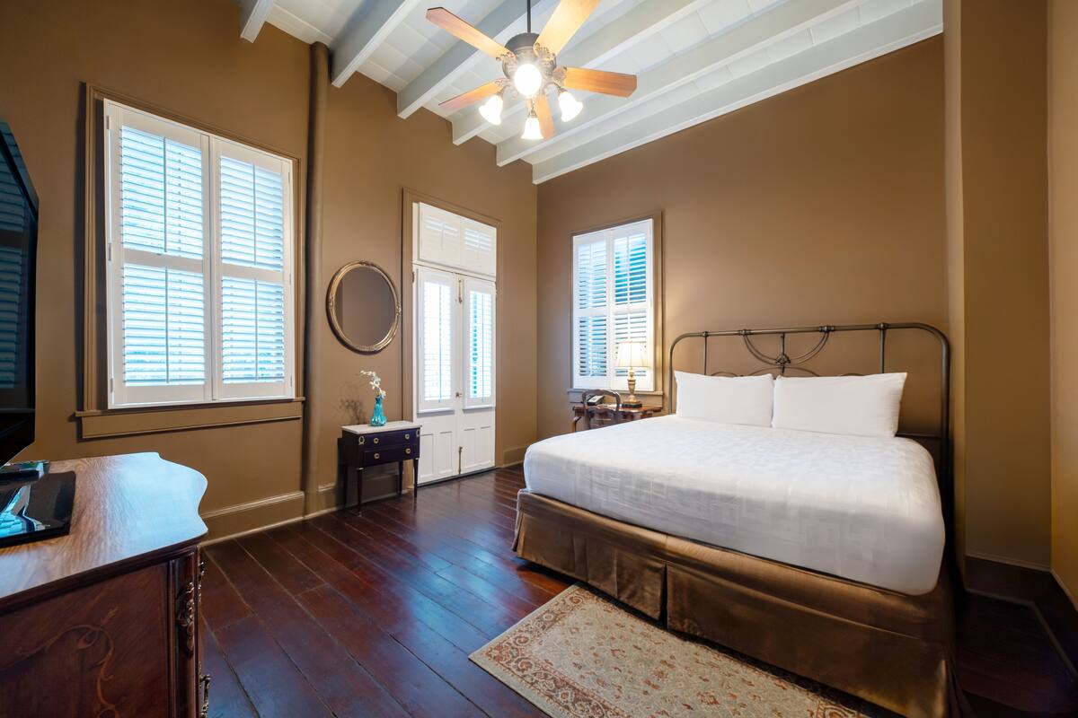 A bedroom with a large bed, wooden flooring, a ceiling fan, and a view of the outside through a window.