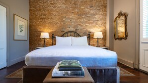 A bedroom with a brick wall, a bed with white linens, two bedside lamps, a mirror, and a stack of books on an ottoman. - Audubon Cottages (New Orleans)