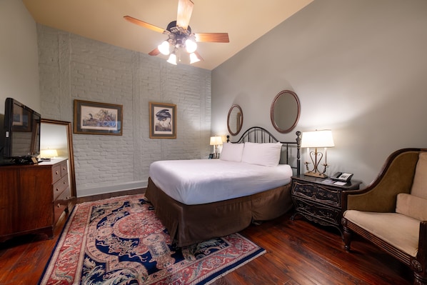 A bedroom with a bed, a wooden dresser, a chair, a nightstand with a lamp, and a ceiling fan. - Audubon Cottages (New Orleans)