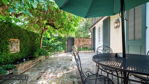 A patio with a brick floor, metal table and chairs, a green umbrella, and a brick wall with a door. - Audubon Cottages (New Orleans)