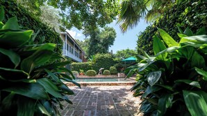 A brick pathway leading to a pool surrounded by lush greenery and a house with a porch. - Audubon Cottages (New Orleans)