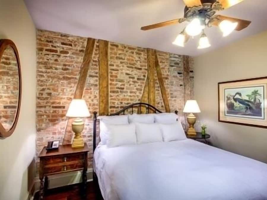 Un dormitorio con pared de ladrillo, una cama con ropa de cama blanca, dos lámparas de noche, un ventilador de techo y un cuadro en la pared.