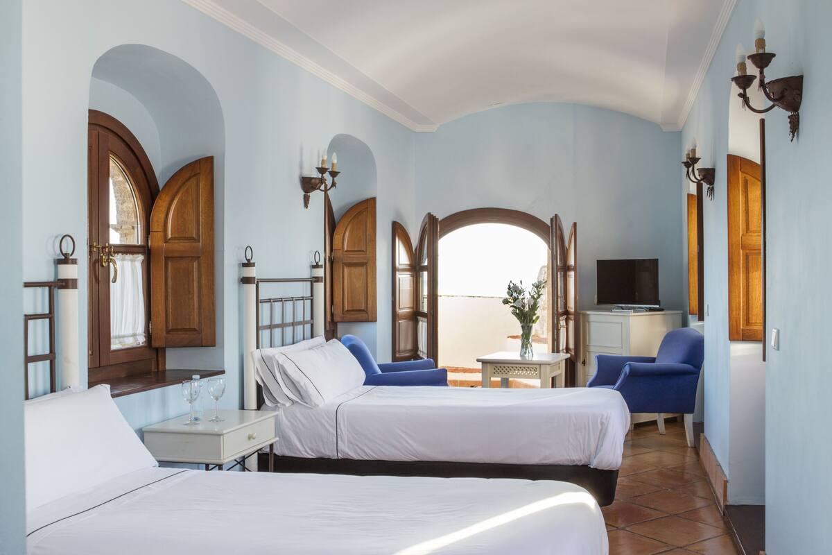 A hotel room with two beds, a TV, a small table, and a view of the sea.