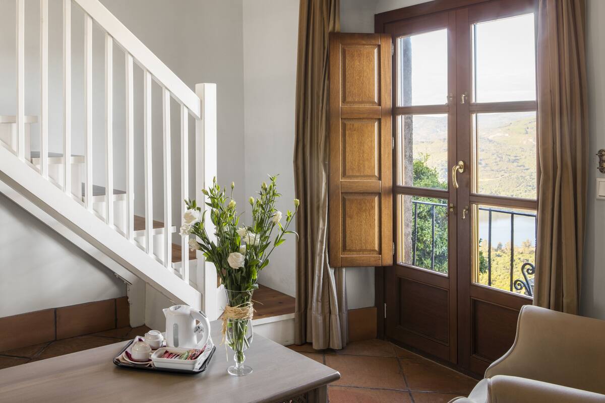 A room with a wooden staircase, a dining table with a vase of flowers, a view of a mountainous landscape through a large window with curtains.