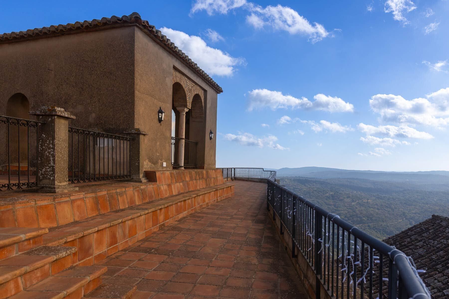 Un edificio de piedra con tejado de tejas, una escalera que conduce a un balcón y una barandilla metálica con vistas a un paisaje montañoso.