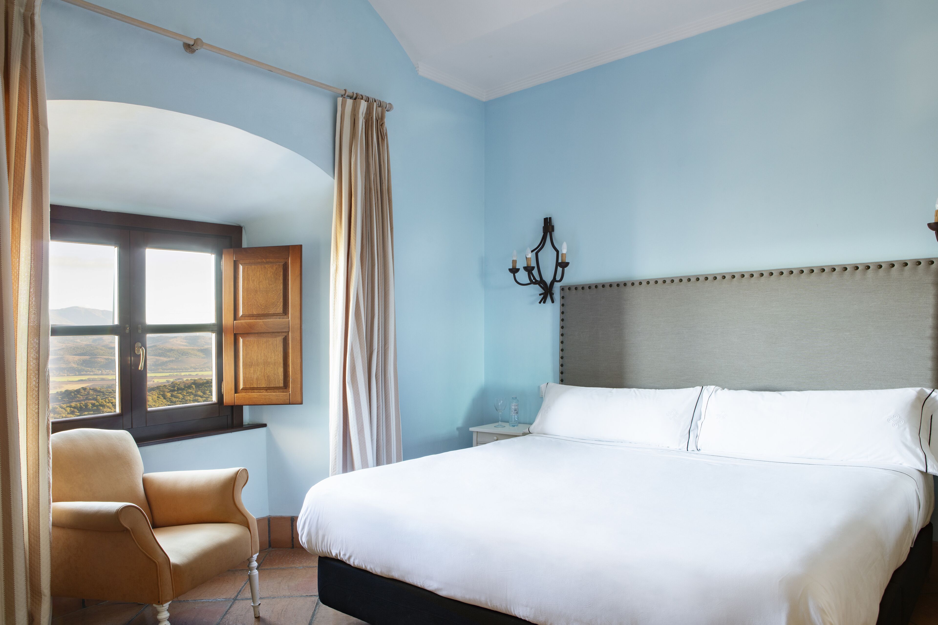 A bedroom with a large bed, a chair, a nightstand, a window with curtains, and a chandelier.