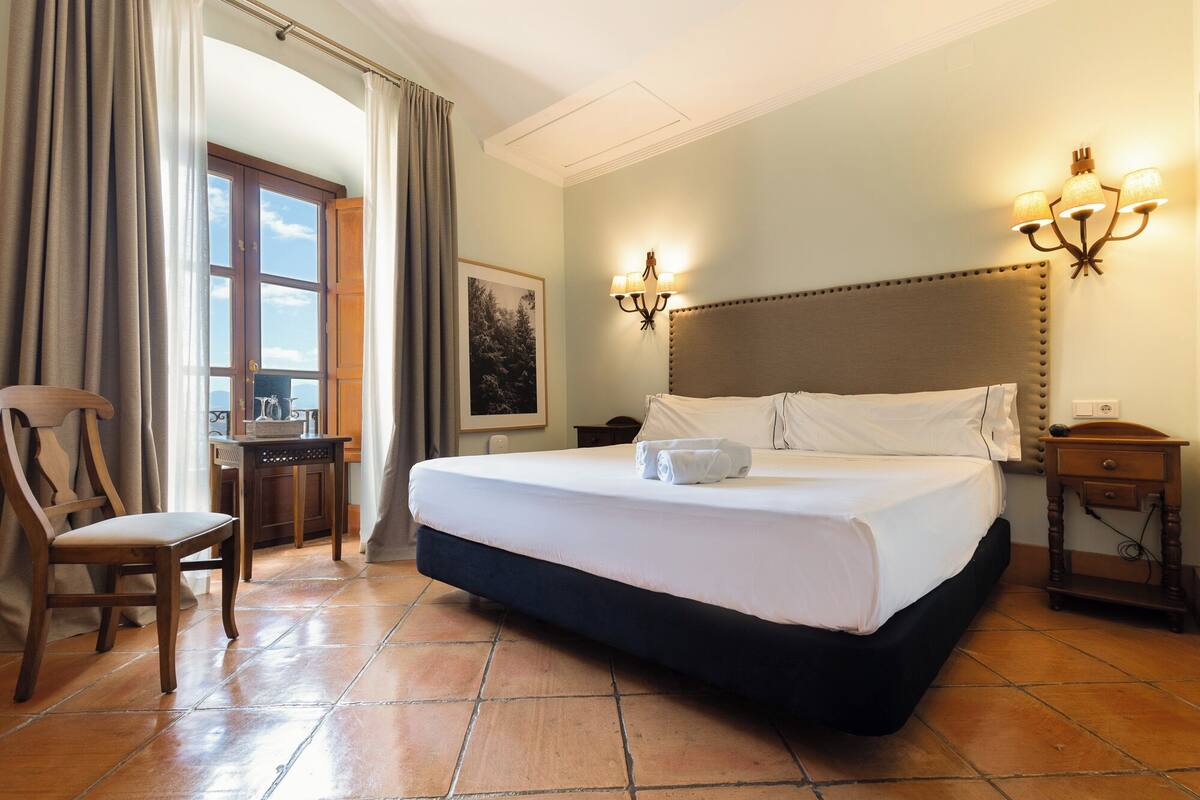 A hotel room with a large bed, a chair, a desk, and a window with curtains.