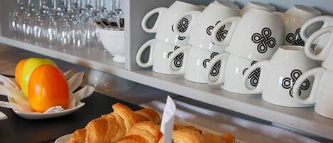A breakfast buffet with croissants, orange slices, and white mugs with a black design.