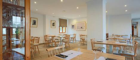 A spacious dining area with multiple tables and chairs, wooden flooring, and framed artwork on the walls.