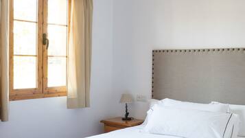 A bedroom with a bed, a window with curtains, a bedside table with a lamp, and a wall socket.