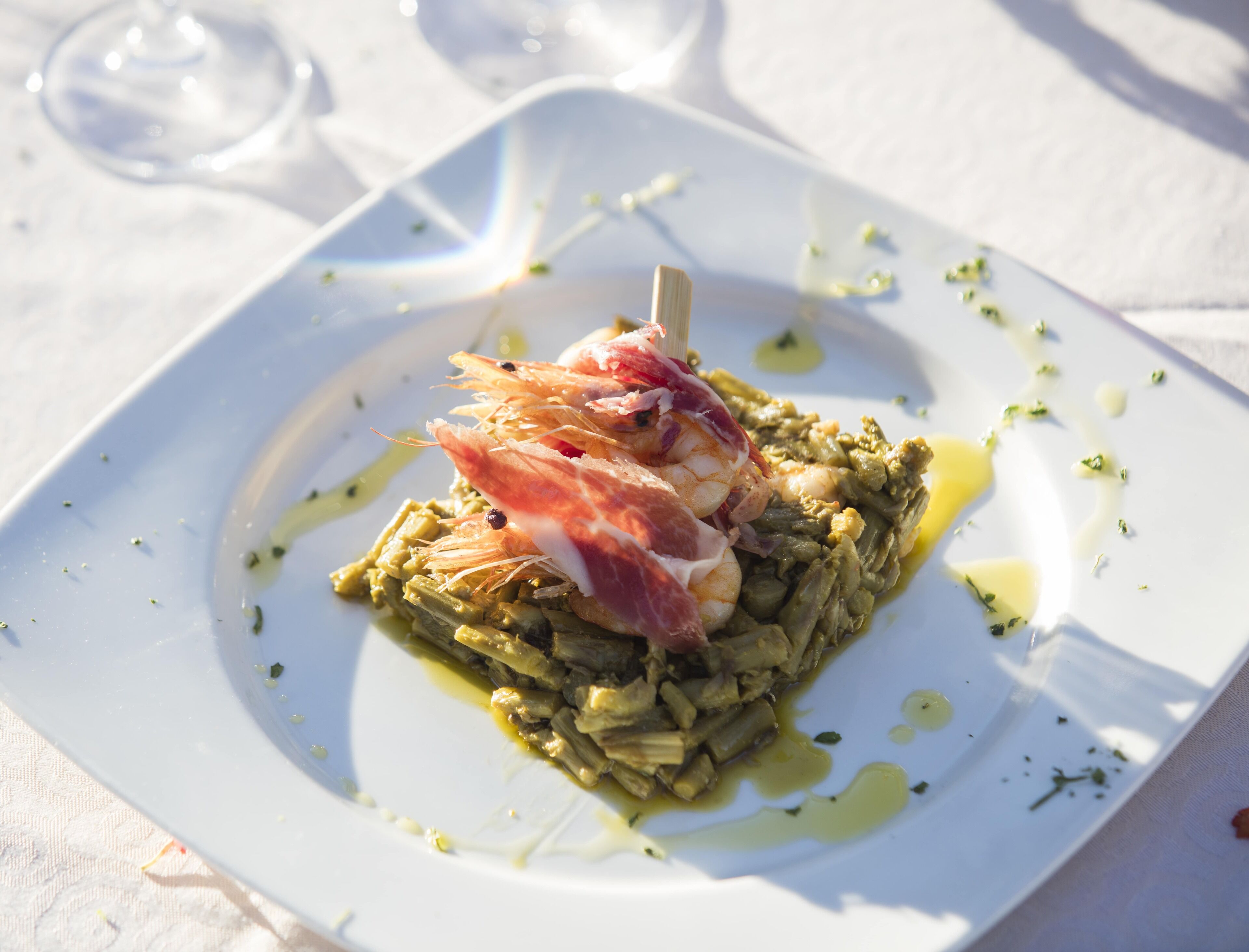 Un plato cuadrado blanco con un guiso de verduras verdes, camarones y un trozo de carne, posiblemente jamón serrano, sobre un mantel blanco.