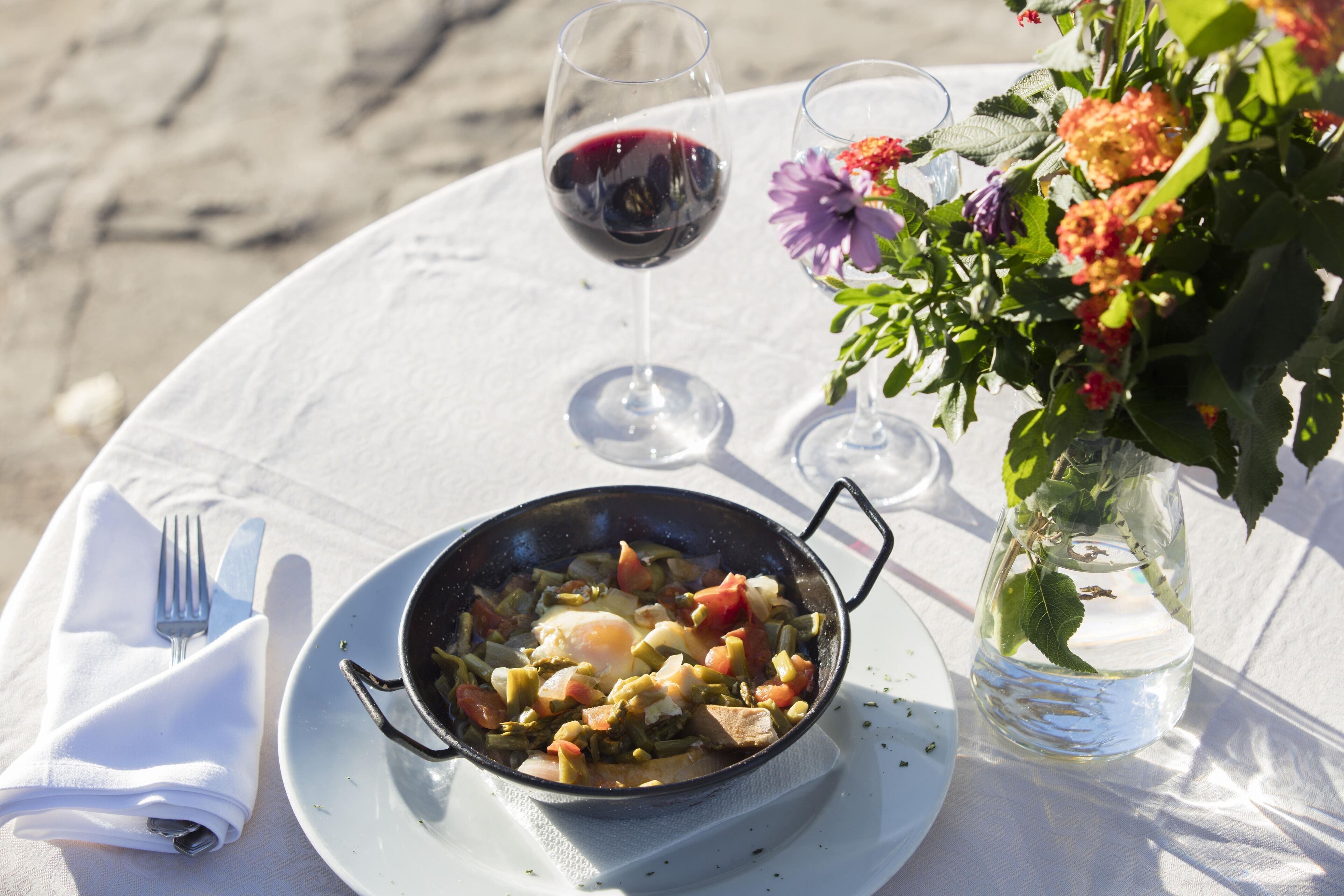 Una mesa servida con comida, copas de vino y un florero con flores.