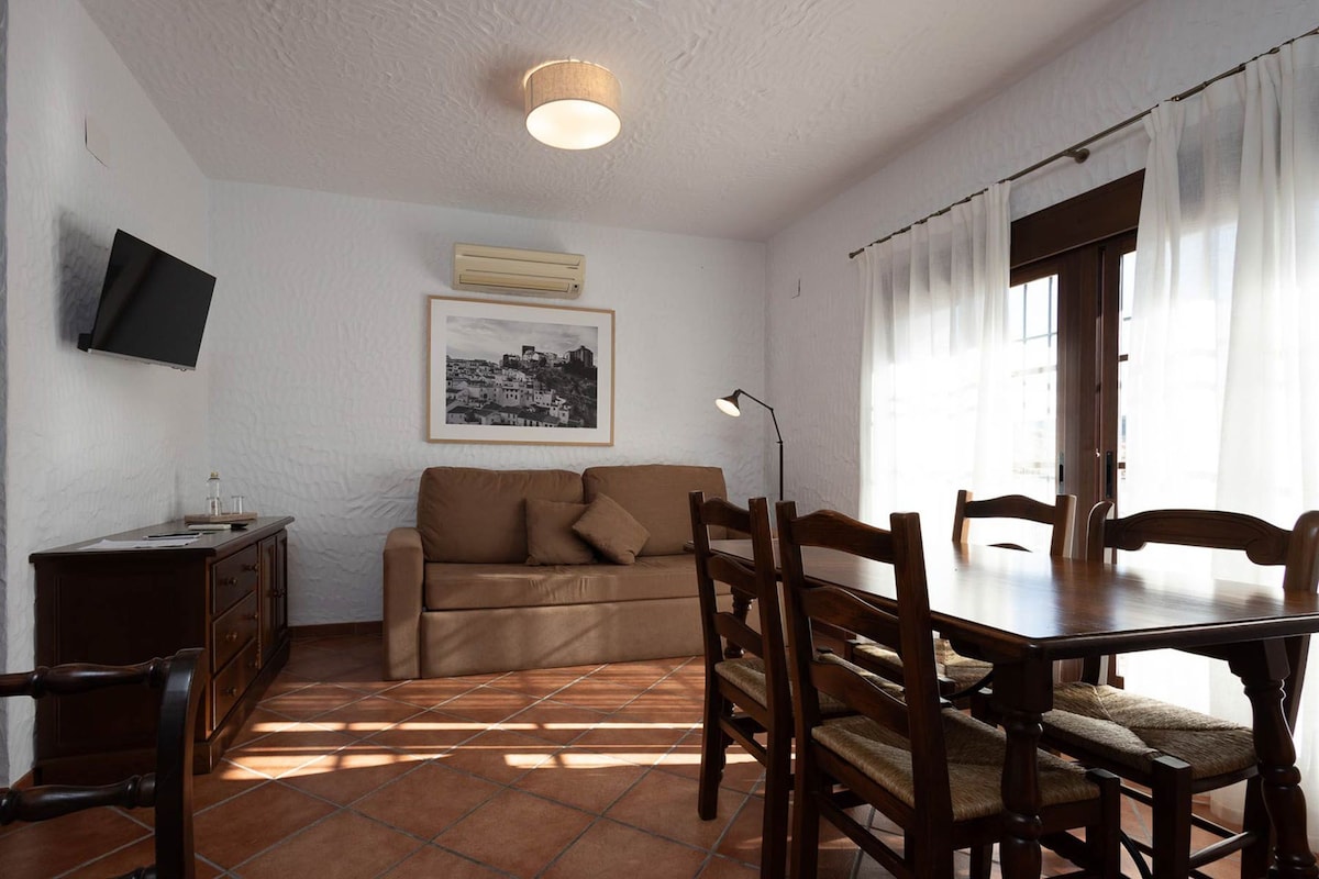 Une pièce avec un canapé marron, une table à manger en bois et des chaises, un téléviseur à écran plat fixé au mur et une fenêtre avec des rideaux.