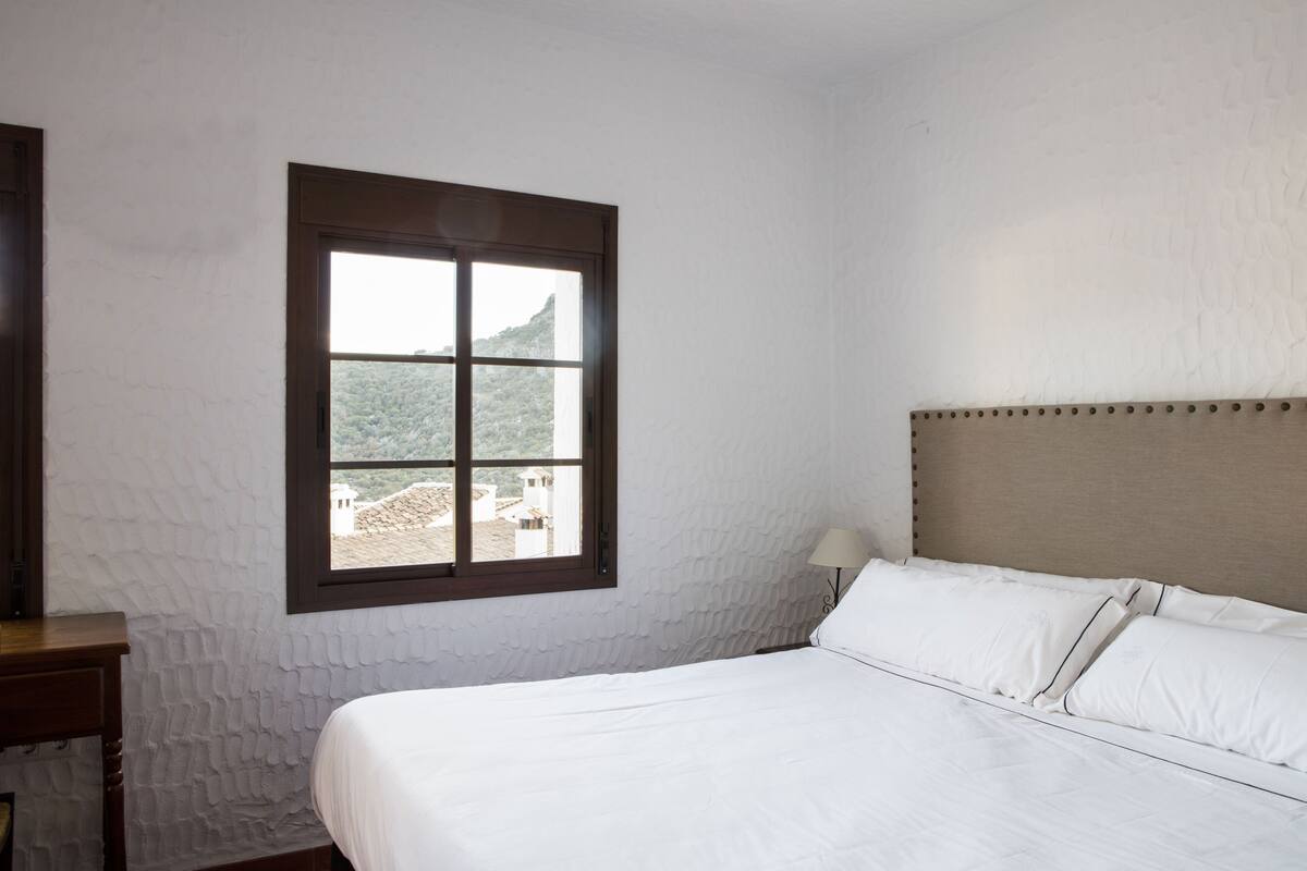A bedroom with a bed, a window with a view of buildings, and a wooden nightstand.