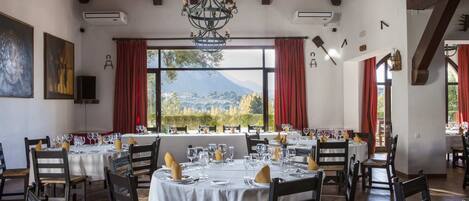 Comedor con mesas dispuestas para una comida, ventanas amplias con vistas a la montaña y vigas de madera en el techo.