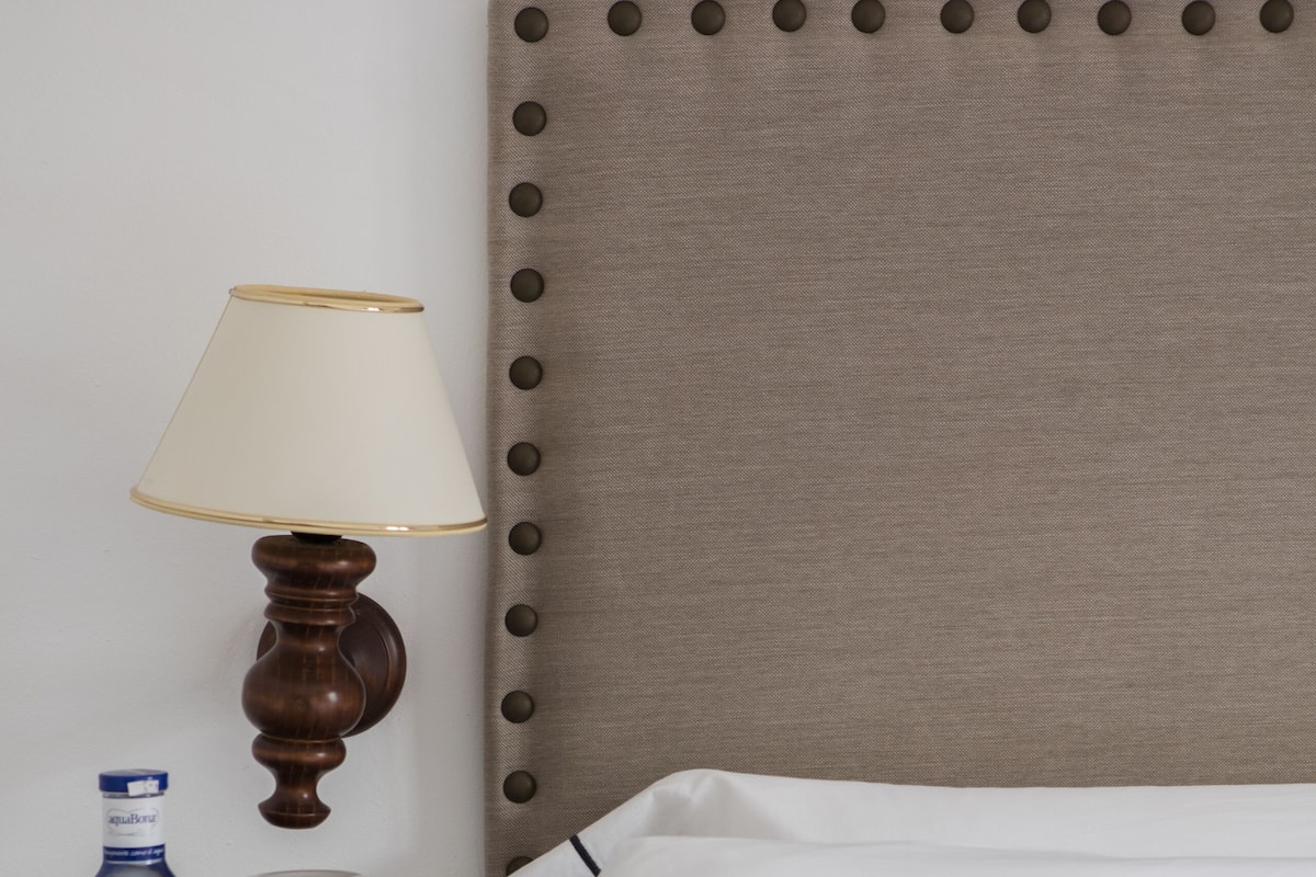 A neatly made bed with white linens, a wooden nightstand with a lamp, a bottle of water, and a wine glass.