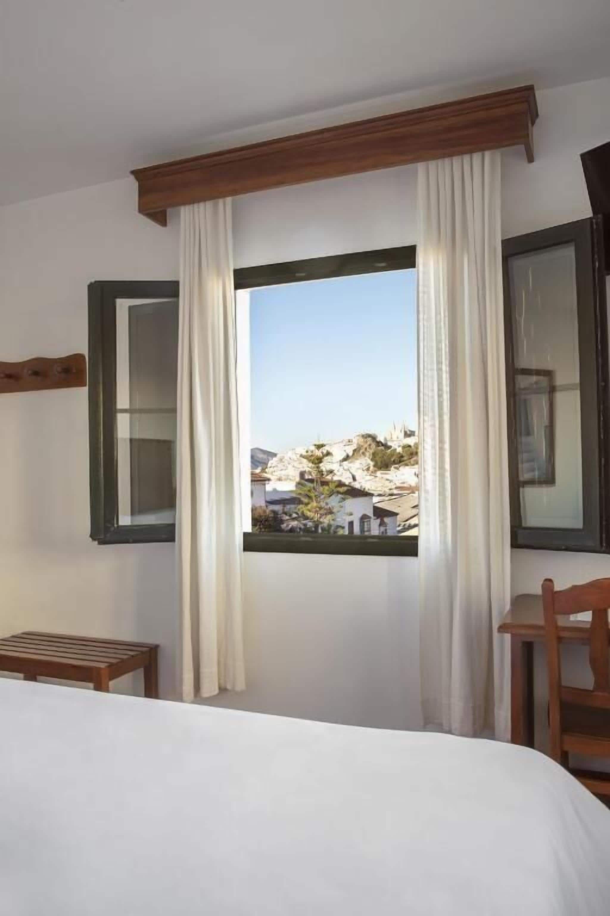 Habitación de hotel con una cama, un banco de madera, una silla y una ventana con cortinas.