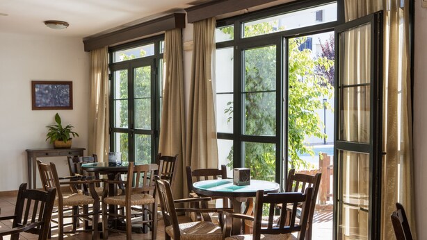 A dining area with wooden chairs and tables, large windows with curtains, and a potted plant on a small table. - TUGASA Sierra Y Cal (Olvera)