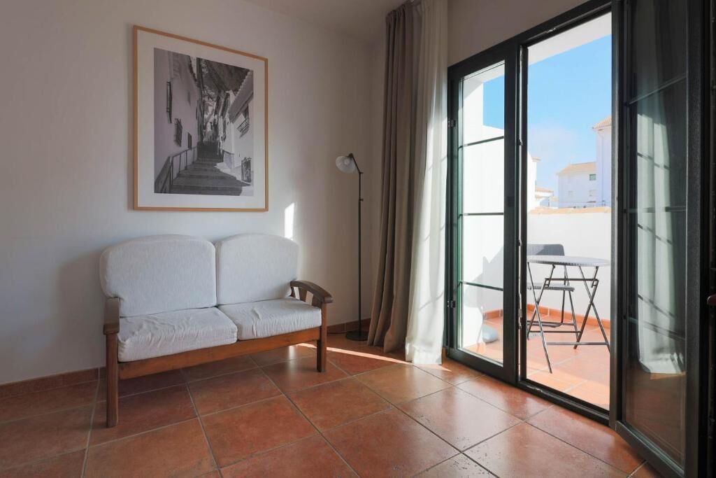 A room with a white sofa, a framed picture on the wall, a floor lamp, and a sliding glass door leading to an outdoor area with a table and chairs.