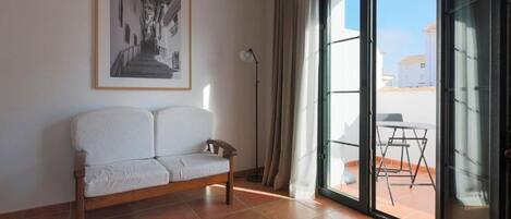 A room with a white sofa, a framed picture on the wall, a floor lamp, and a sliding glass door leading to an outdoor area with a table and chairs.
