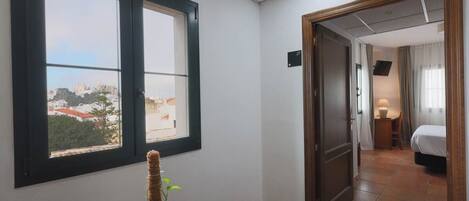 A hotel room with a view of a cityscape through large windows, a wooden door leading to another room, and a small bedside table with a lamp.