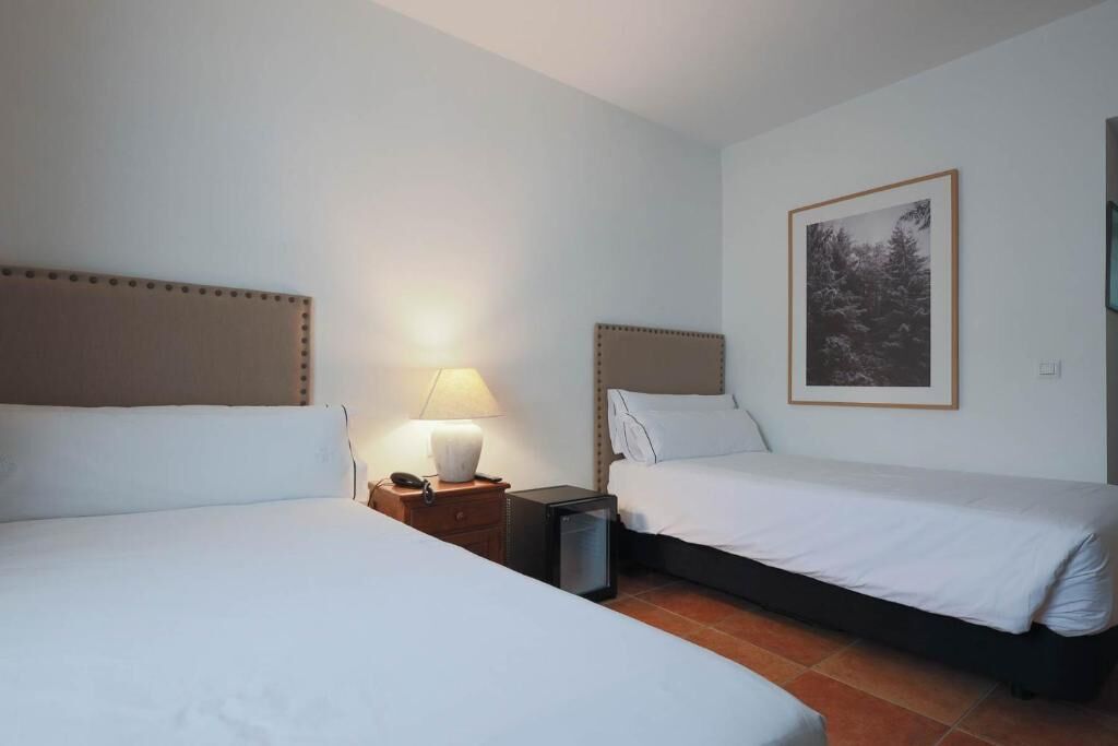 Habitación de hotel con dos camas, una mesita de noche con lámpara, un pequeño cubo de basura y un cuadro en la pared.