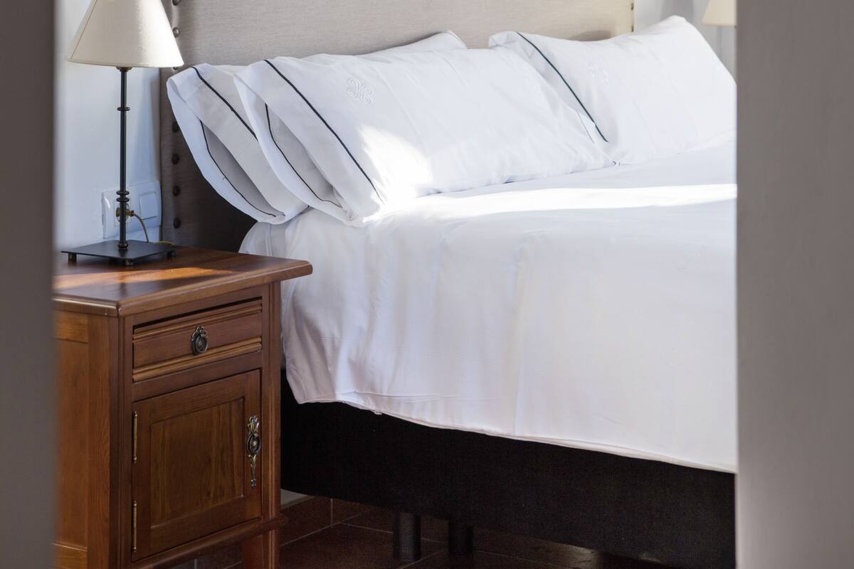 A neatly made bed with a headboard, a wooden nightstand with a lamp, and a wall-mounted light fixture.