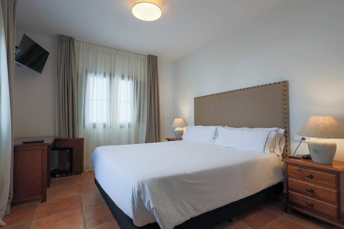A hotel room with a large bed, a TV mounted on the wall, a bedside table with a lamp, and a window with curtains.