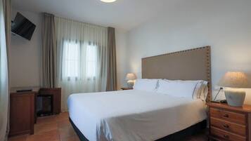 Habitación de hotel con una cama grande, televisor montado en la pared, mesita de noche con lámpara y ventana con cortinas.