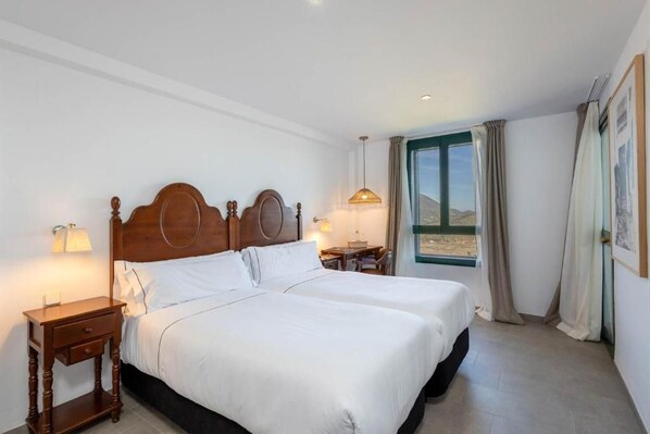A double bed with a wooden headboard, two bedside tables with lamps, a view of the outdoors through a window, and a small table with a chair.