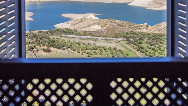 Vista pela janela aberta, com grade de metal, sobre um vale com lago e colinas.