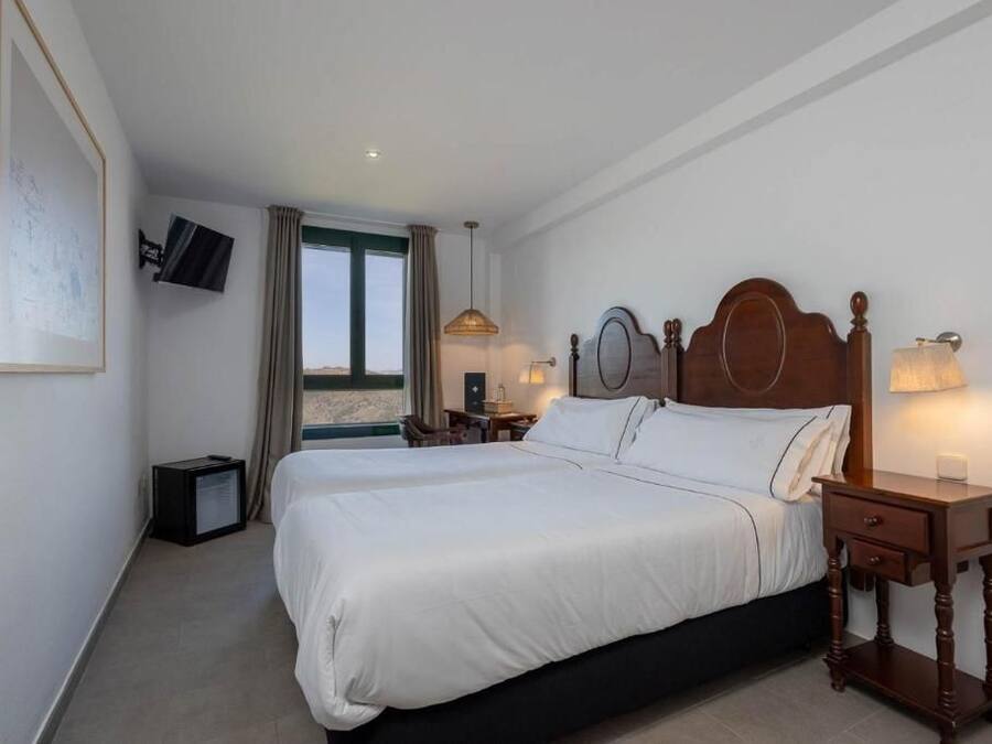 Una habitación de hotel con una cama grande, un televisor montado en la pared, una mesita de noche con lámpara y una ventana con cortinas.