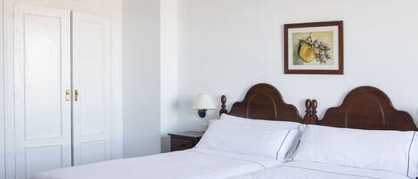 Una cama bien hecha con sábanas blancas, cabecera de madera, mesita de noche con lámpara y un cuadro enmarcado en la pared.