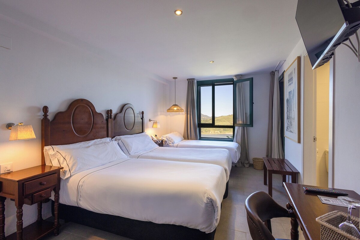 A hotel room with two beds, a desk, a chair, a window with a view, and a lamp.