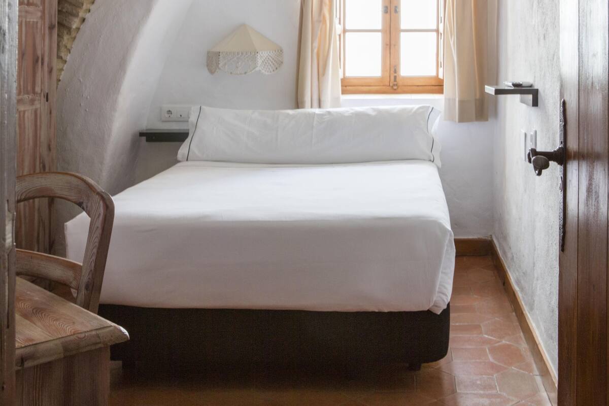 A bedroom with a bed, a window with curtains, a wooden door, and a tiled floor.