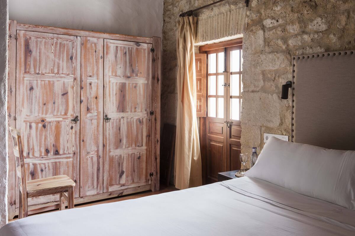 A bedroom with a large wooden wardrobe, a bed with white linens, and a stone wall.