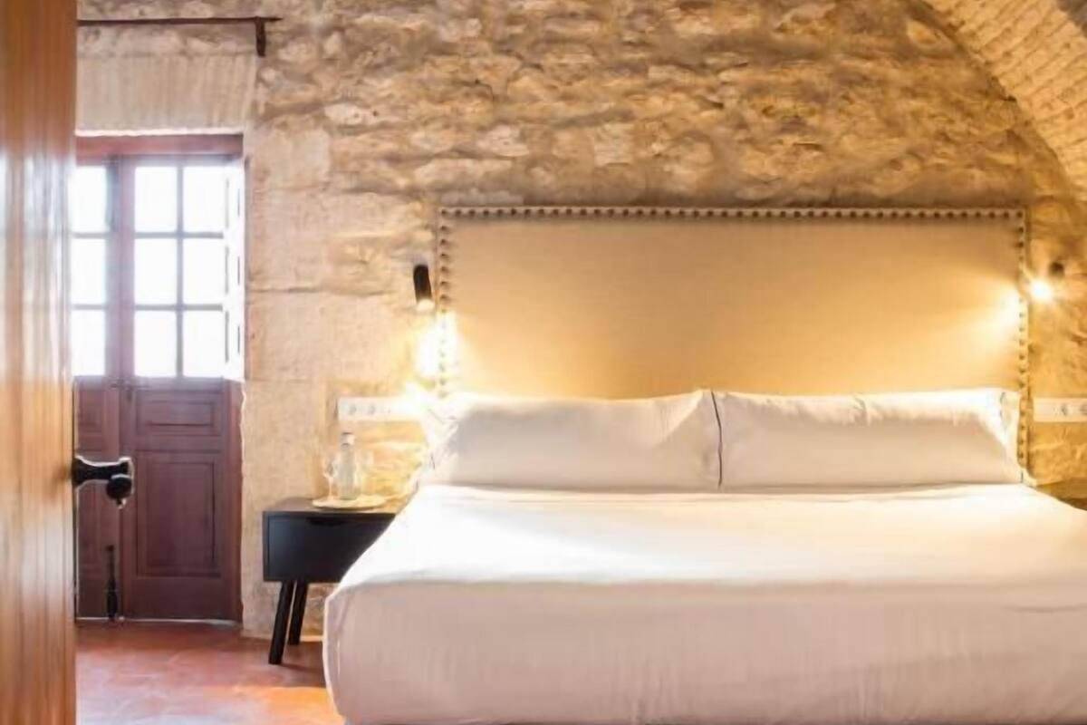 A bedroom with a stone vaulted ceiling, a large bed with white linens, a bedside table with a lamp, and a wooden door.